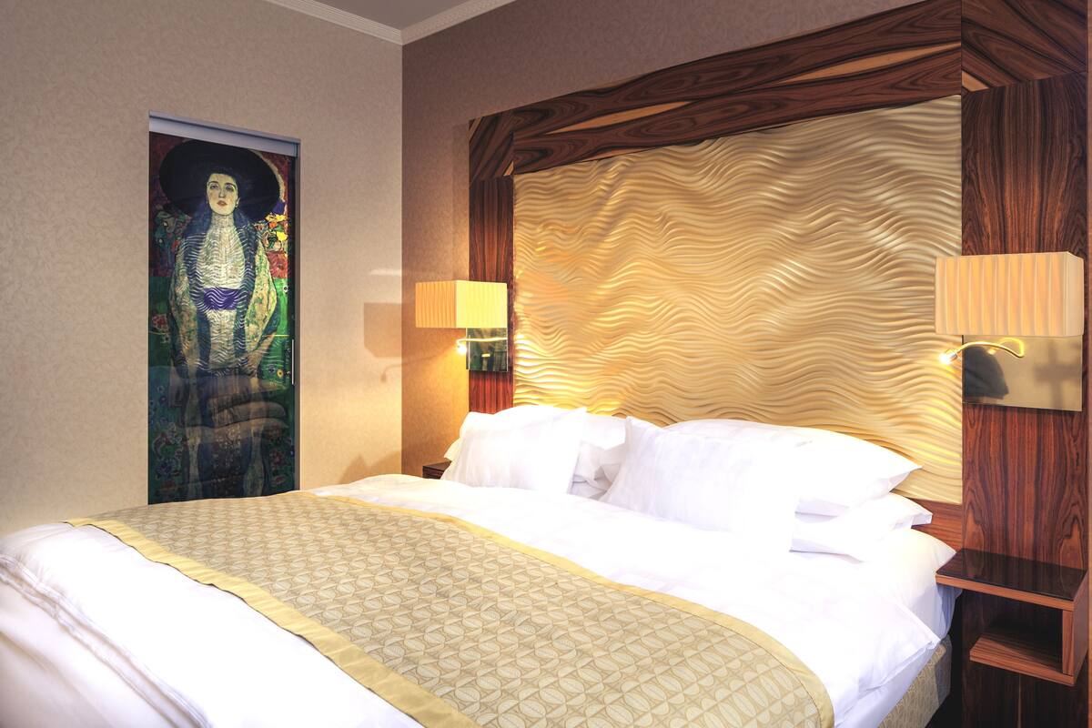 Quarto de hotel com uma cama grande, cabeceira de madeira, luminária acoplada à parede e uma obra de arte emoldurada.