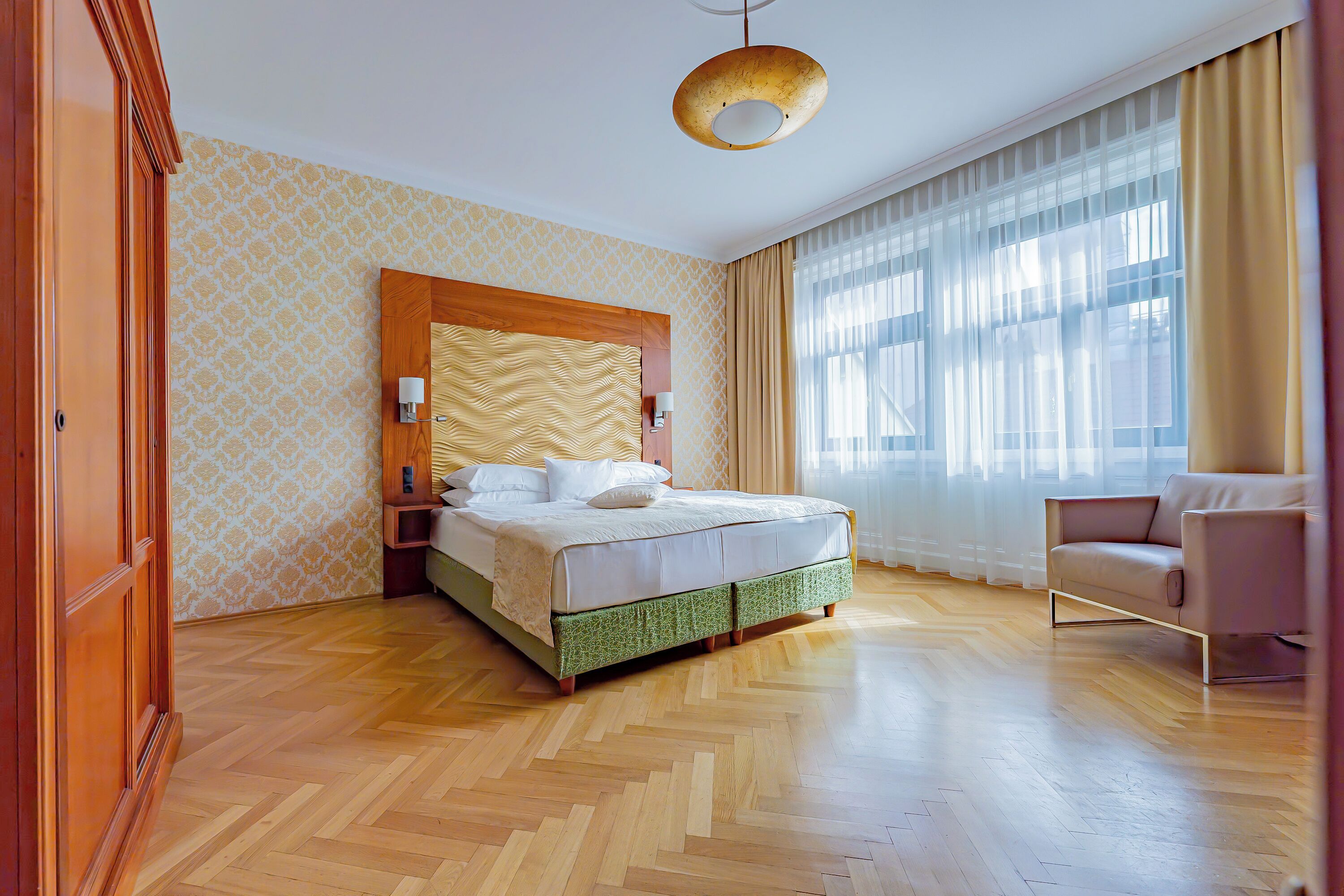 Habitación de hotel con suelo de madera en patrón de espiga, una cama grande con cabecera de madera, un sofá y un ventanal con cortinas traslúcidas.
