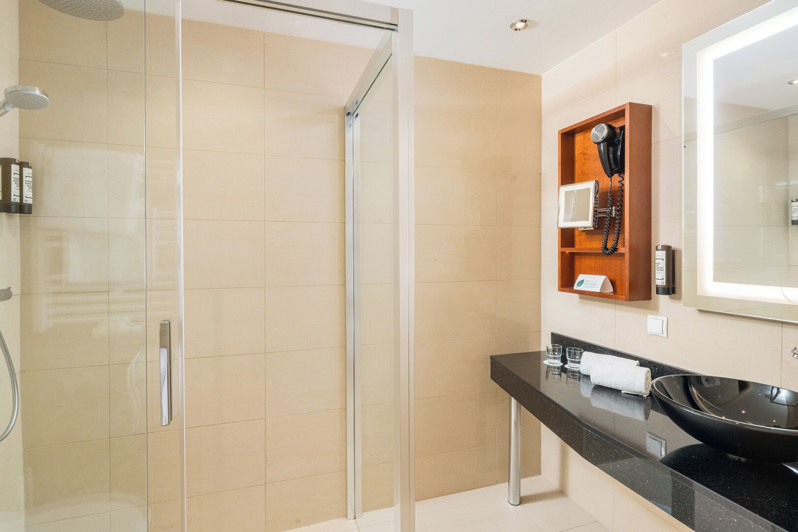 Een moderne badkamer met een douchecabine van glas, een zwarte wastafel en een houten badkamermeubel met een haardroger en spiegel.