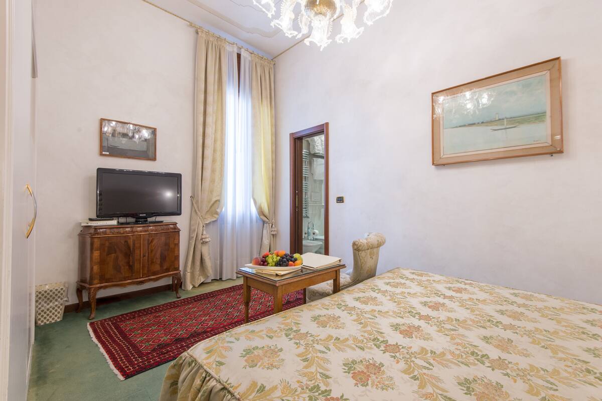 Ein Schlafzimmer mit einem Bett, einem Fernseher auf einer Holzkabine, einer Obstschale auf einem kleinen Tisch und einem Bild an der Wand.