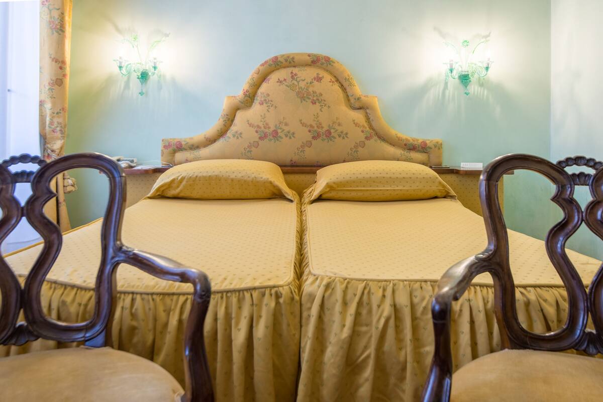 Ein Doppelbett mit floralem Kopfstück und zwei Wandleuchten, flankiert von verzierten Holzstühlen.