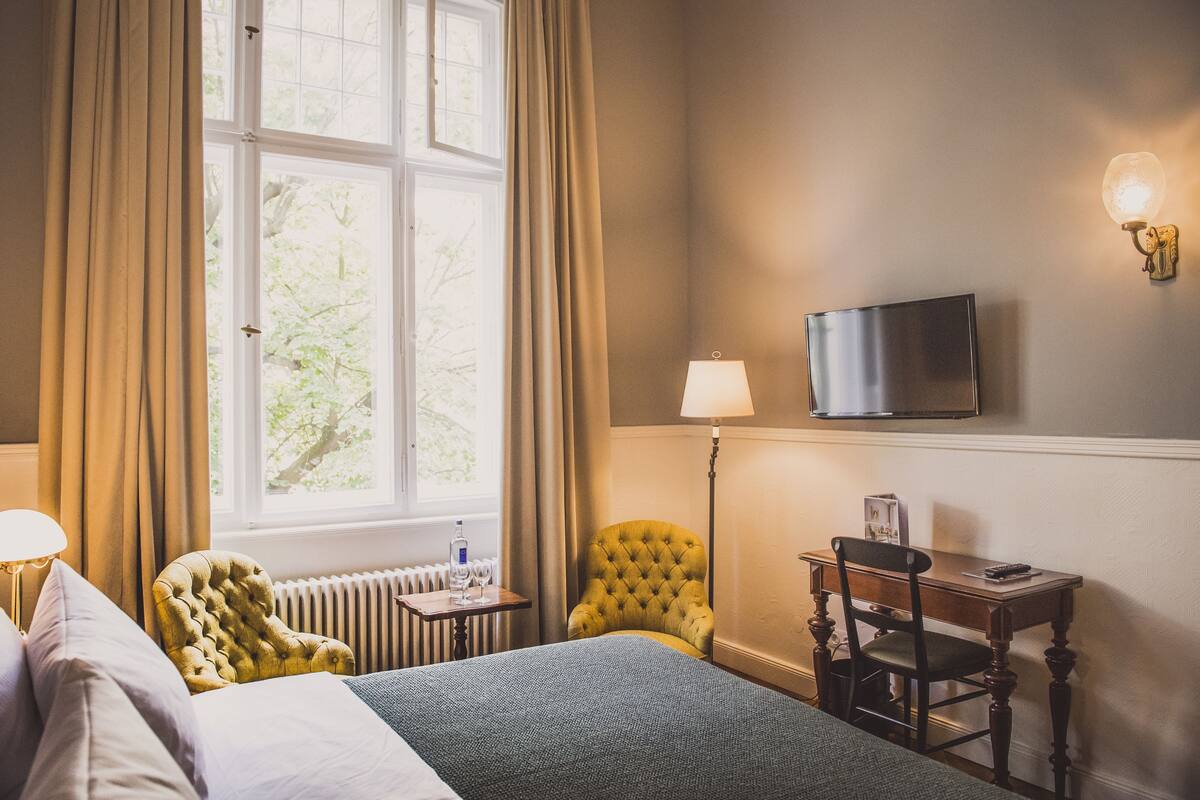 A hotel room with a bed, a yellow armchair, a desk with a chair, a lamp, a television, and a window with curtains.