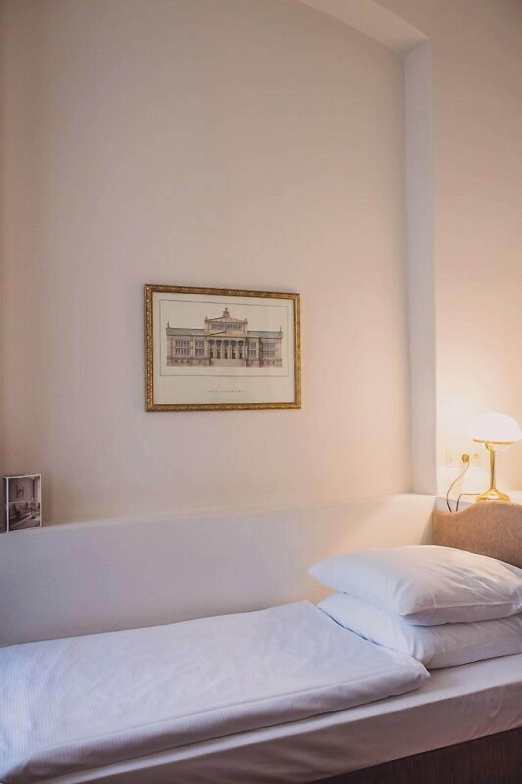 A single bed with white bedding in a room with a framed picture on the wall and a bedside lamp.