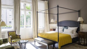 A four-poster bed with a yellow bedspread, a bench at the foot of the bed, a chair with a patterned upholstery, and a small table. - Henri Hotel Berlin (Berlin)