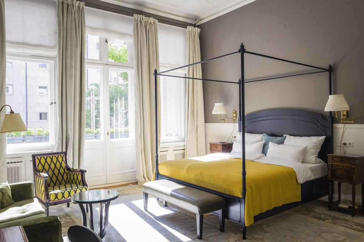 A four-poster bed with a yellow bedspread, a bench at the foot of the bed, a chair with a patterned upholstery, and a small table.