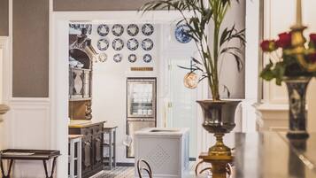 A dining area with wooden chairs, a table, and a vase with flowers. There is a kitchen in the background with various appliances and a refrigerator.