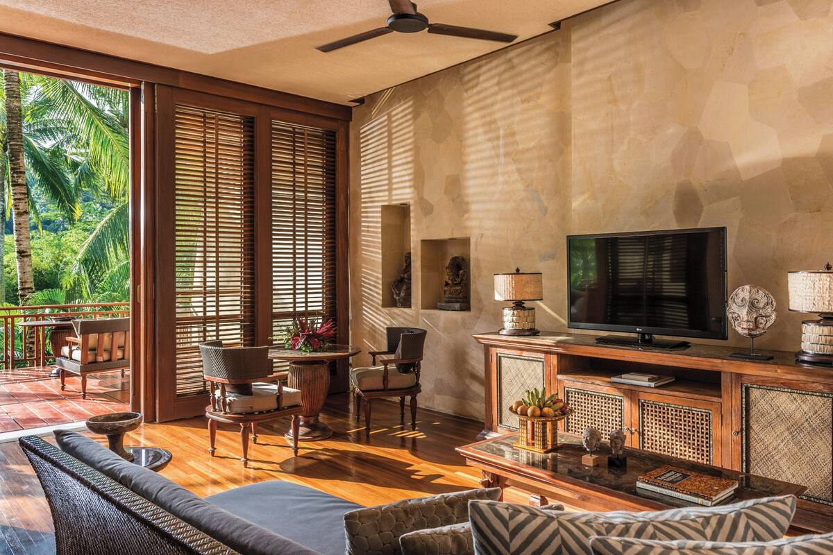 A spacious living area with a large TV, wooden furniture, and a balcony with a view of lush greenery.