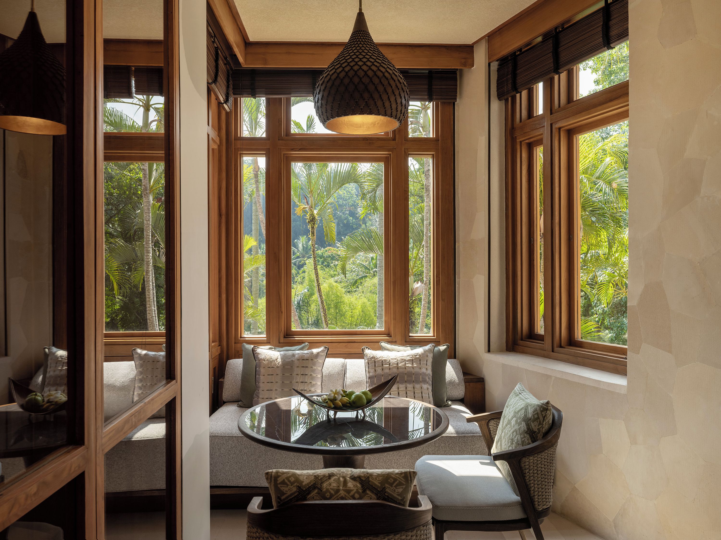 Una habitación con una mesa redonda de vidrio, asientos acolchados, ventanas amplias con vistas a la vegetación y una lámpara colgante.
