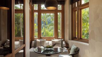 Una habitación con una mesa redonda de vidrio, asientos acolchados, ventanas amplias con vistas a la vegetación y una lámpara colgante.