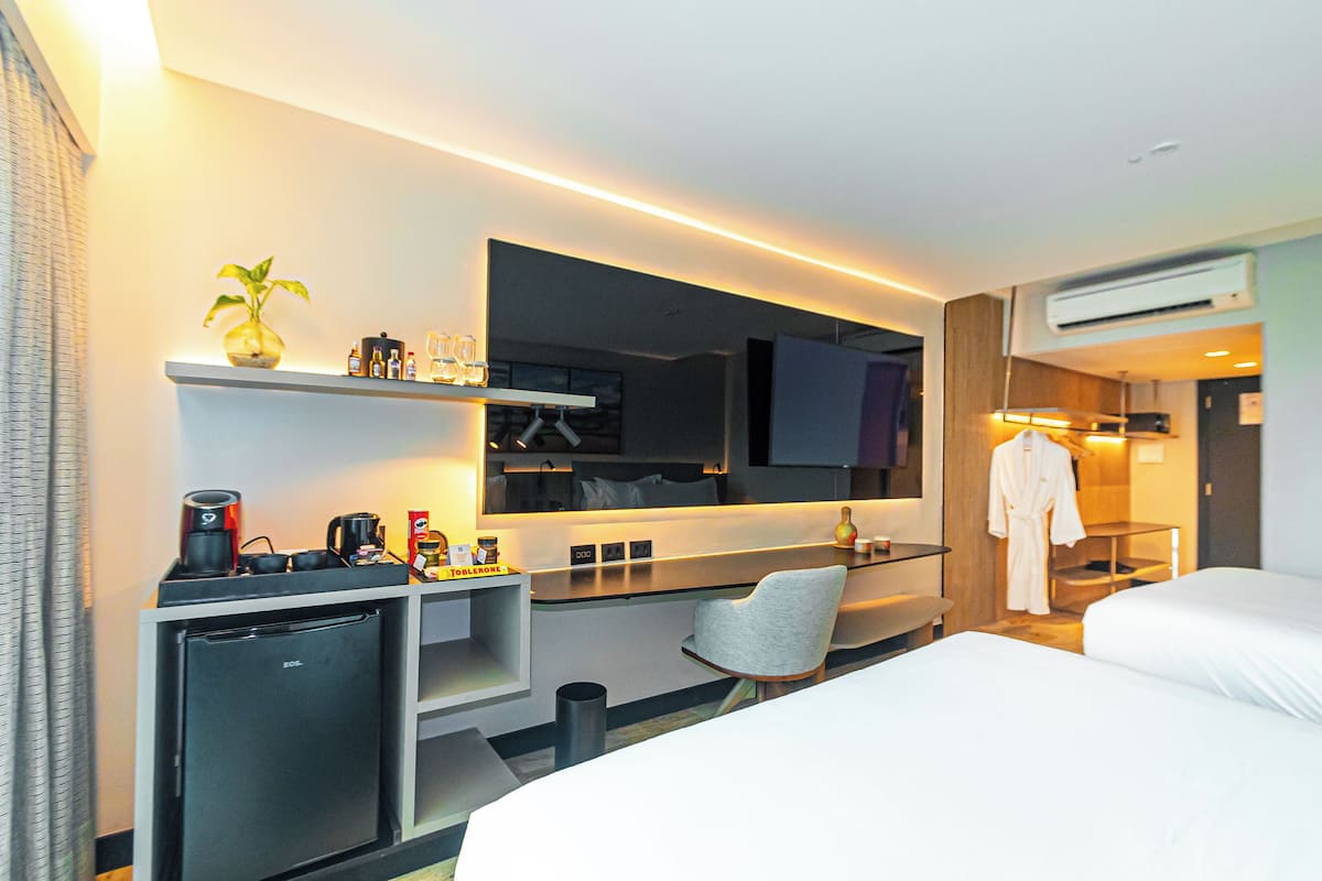 A modern hotel room with a bed, a desk with a computer, a chair, a small fridge, a TV, and a robe hanging on a rack.