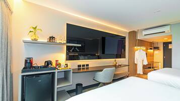 Habitación de hotel moderna con cama, escritorio con ordenador, silla, refrigerador pequeño, televisor y bata colgada en un perchero.