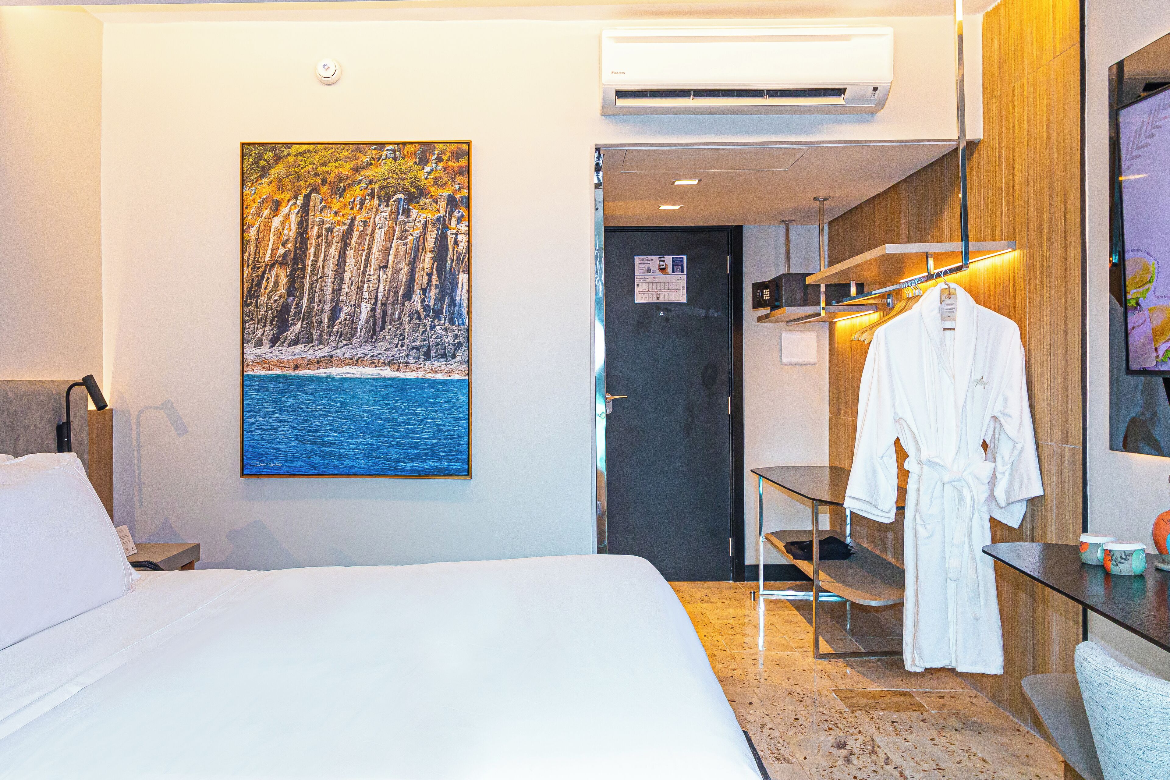 A hotel room with a bed, a painting of a coastal scene, an air conditioner, a robe hanging on a rack, and a small table with a cup and a bowl.