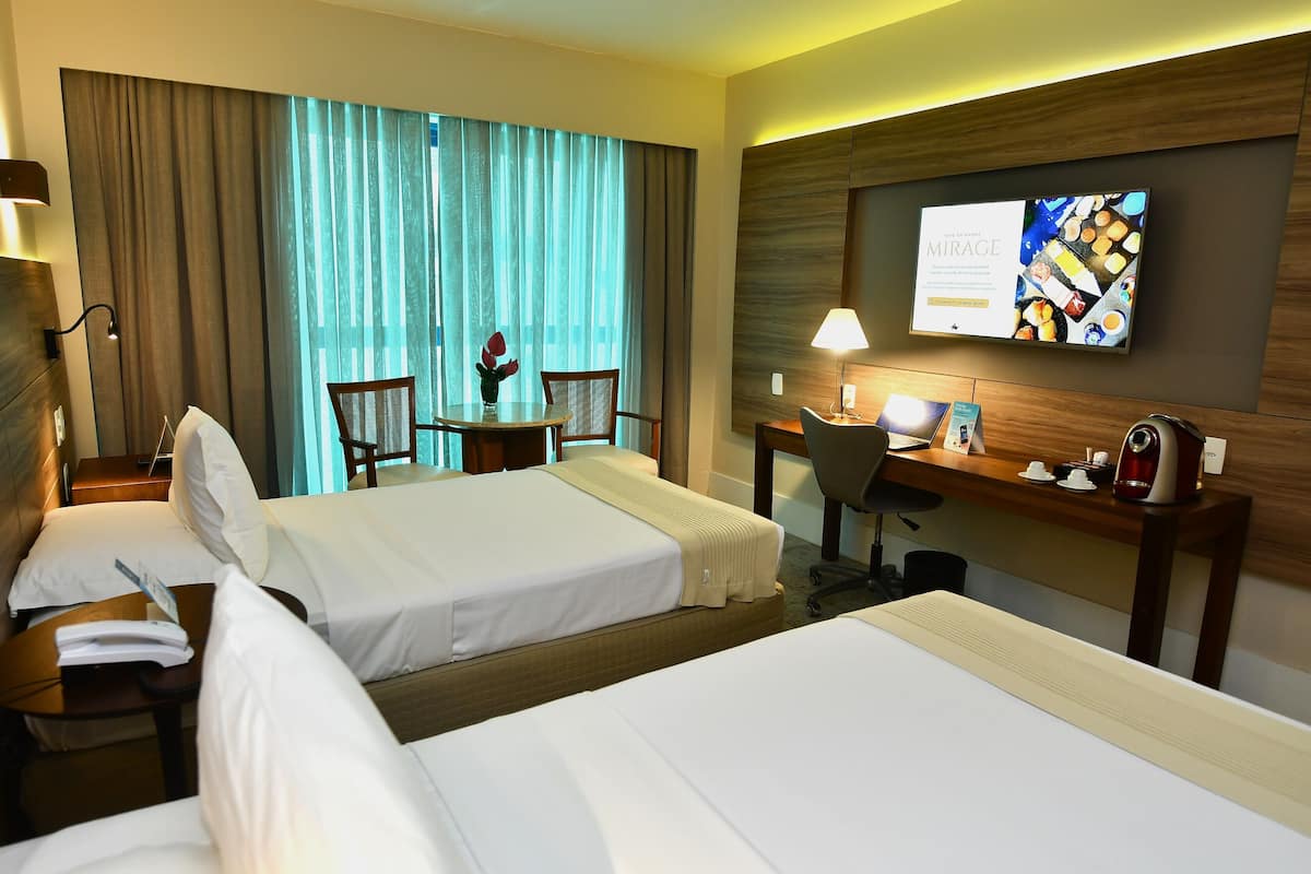 A hotel room with two beds, a desk with a lamp, a chair, a TV on the wall, and a window with curtains.
