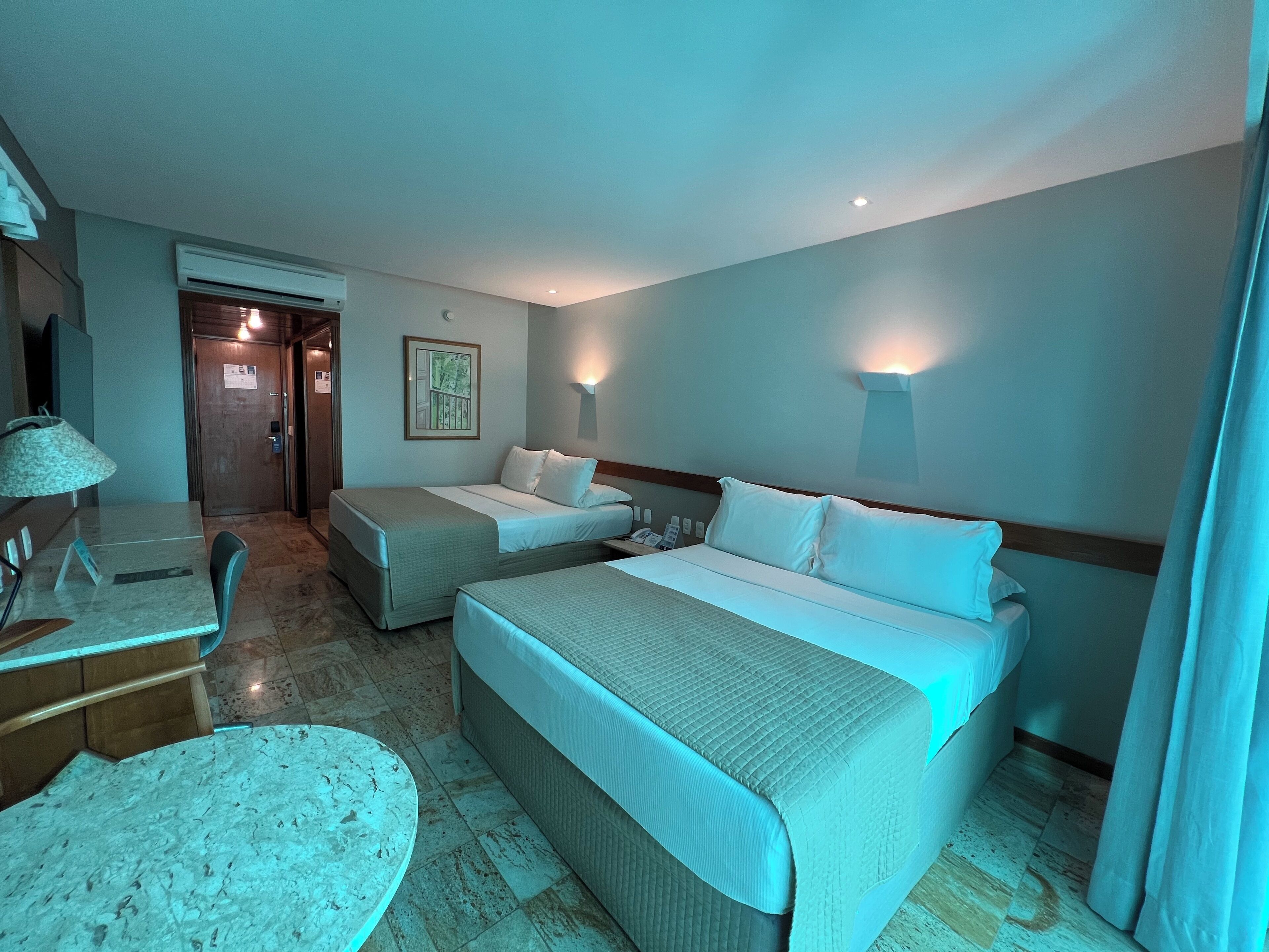 A hotel room with two beds, a desk with a chair, a small table, and a wall-mounted lamp.