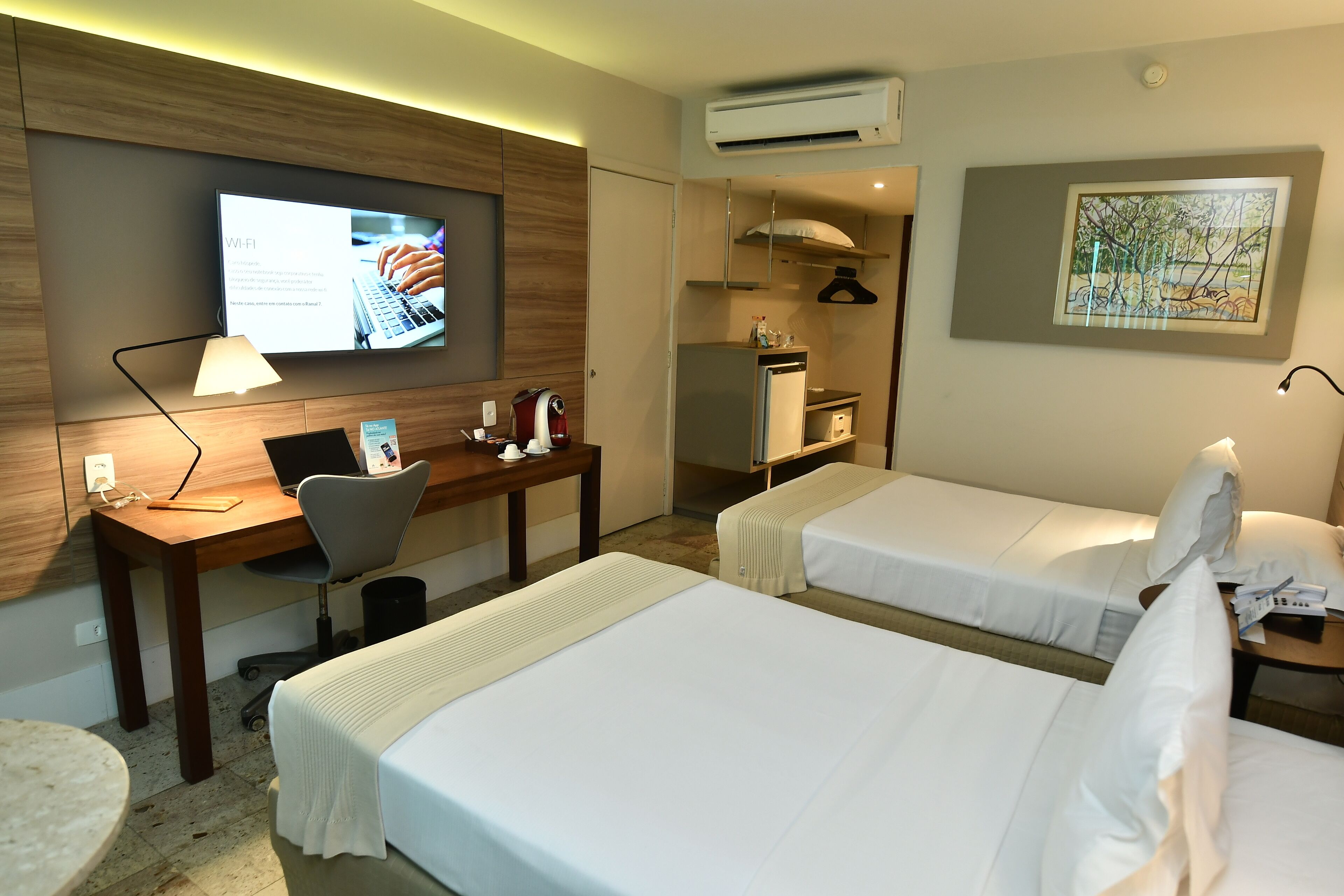 Habitación de hotel con dos camas, un escritorio con una computadora portátil, un televisor mostrando información, una zona de cocina y un cuadro en la pared.