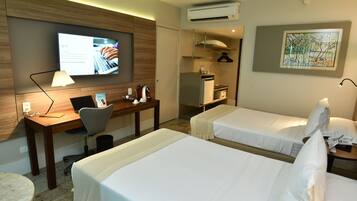 Habitación de hotel con dos camas, un escritorio con una computadora portátil, un televisor mostrando información, una zona de cocina y un cuadro en la pared.