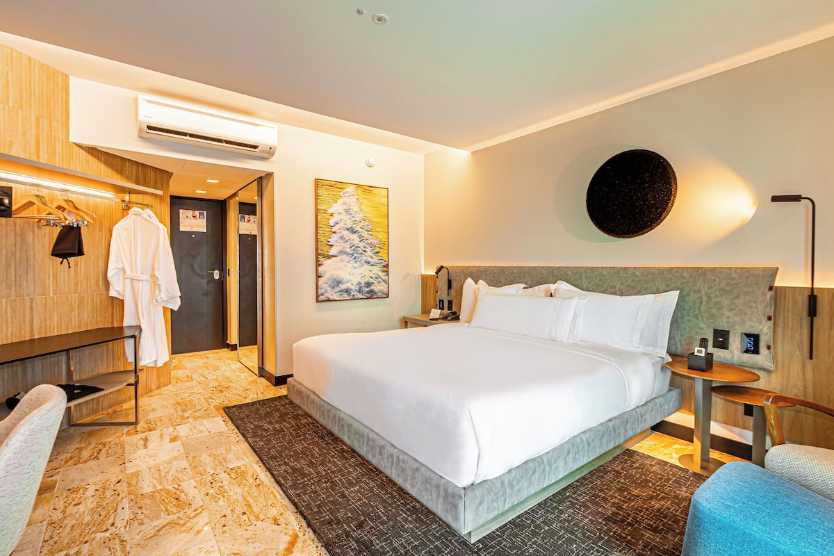 A hotel room with a large bed, a robe rack, a towel rack, a small table with a lamp, and a wall-mounted air conditioner.
