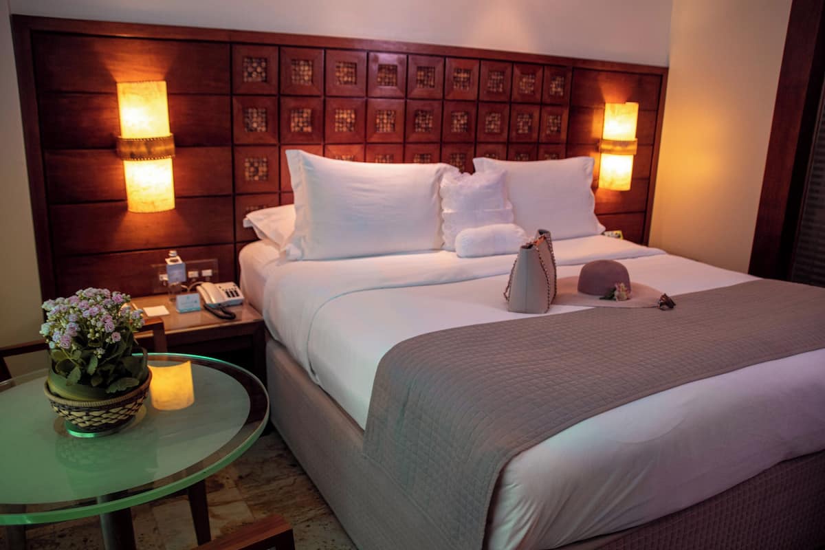 A hotel room with a large bed, two bedside tables, a nightstand, a glass table with a vase of flowers, and a decorative headboard with wall sconces.