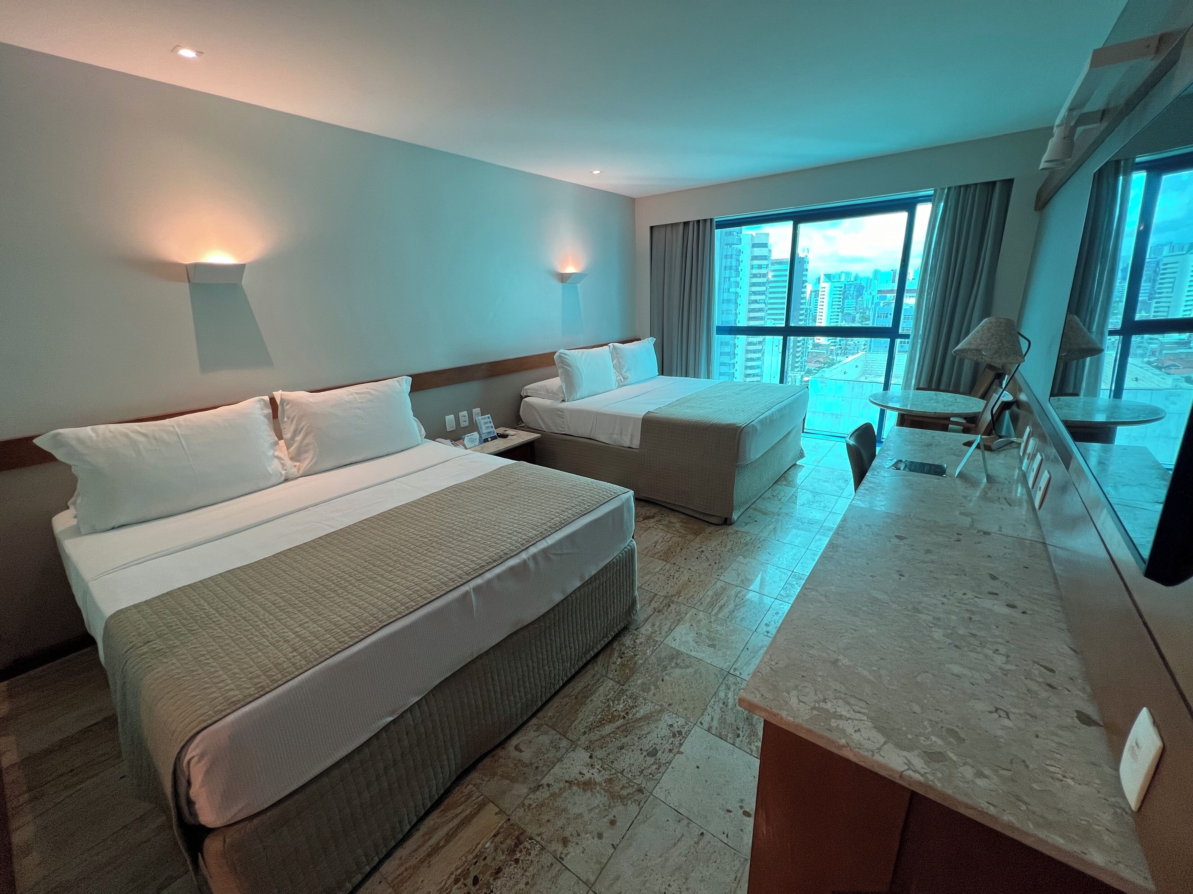 A hotel room with two beds, a desk, and a large window offering a city view.