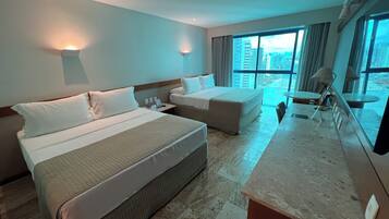 A hotel room with two beds, a desk, and a large window offering a city view.
