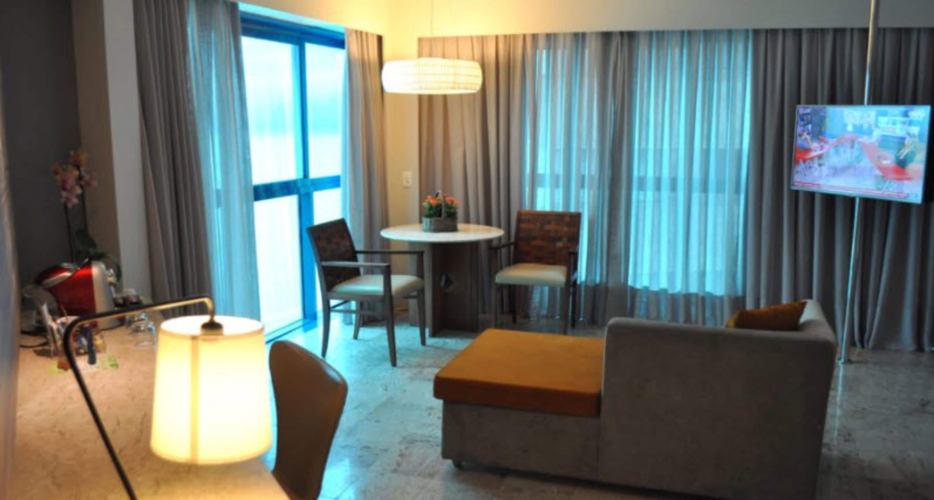A hotel room with a sofa, chairs, a small table, a lamp, a TV, and curtains.