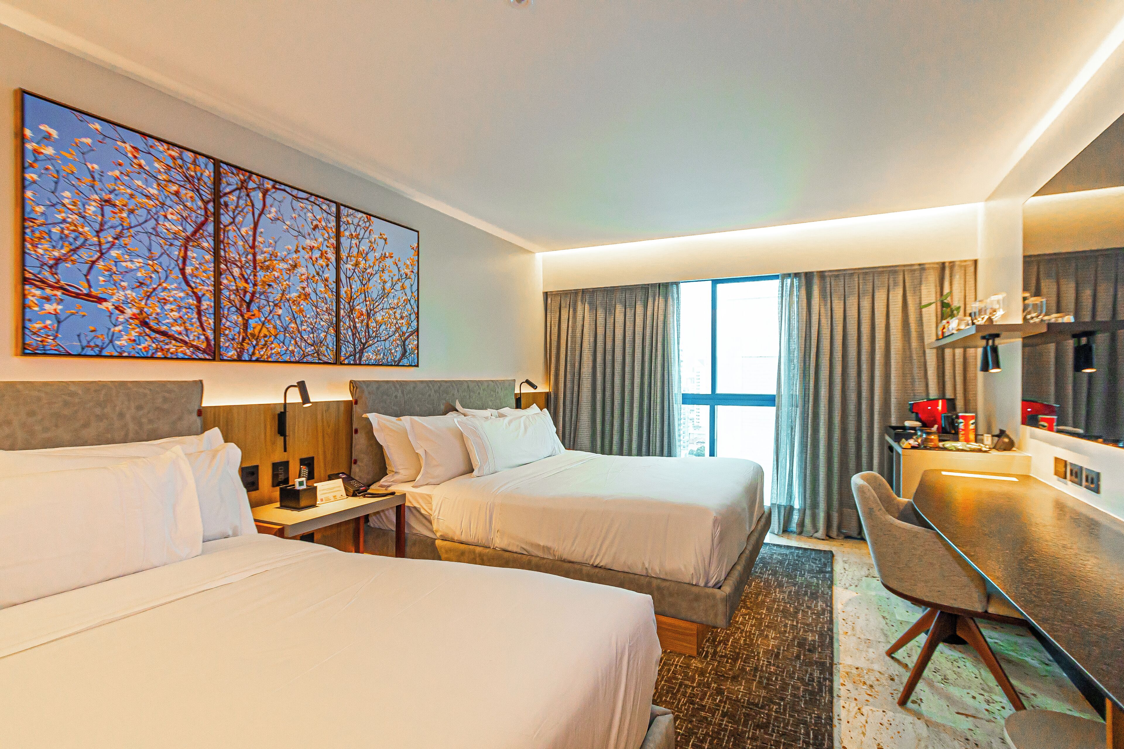 A hotel room with two beds, a desk, a chair, a large window with curtains, and a painting on the wall.