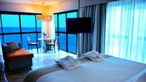 Habitación de hotel con una cama grande, un sofá, una mesa con sillas, un televisor y vistas al océano.