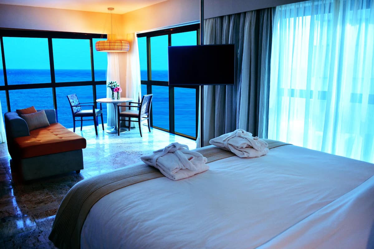 A hotel room with a large bed, a sofa, a table with chairs, a TV, and a view of the ocean.