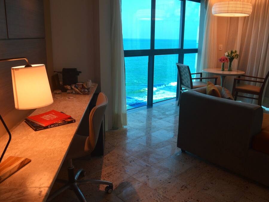 Quarto de hotel com uma mesa, um sofá, vista para o oceano e uma luminária.