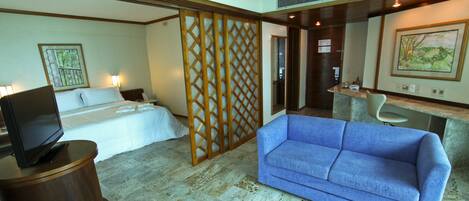 Habitación de hotel con cama, sofá azul, televisor sobre un soporte de madera, escritorio con una silla y un panel decorativo.