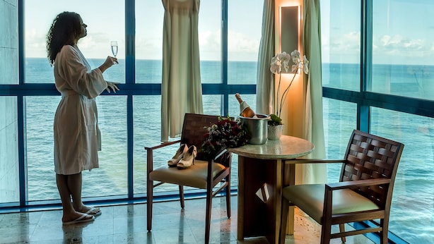 Una habitación con un ventanal grande con vista al mar, una mesa con sillas, un vestido colgado y un jarrón con flores.