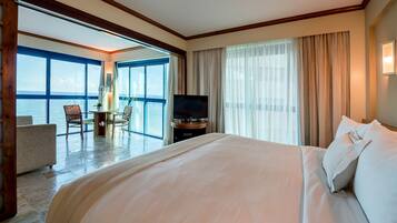 Habitación de hotel con una cama grande, televisión, escritorio con silla y vistas al mar a través de ventanas amplias.