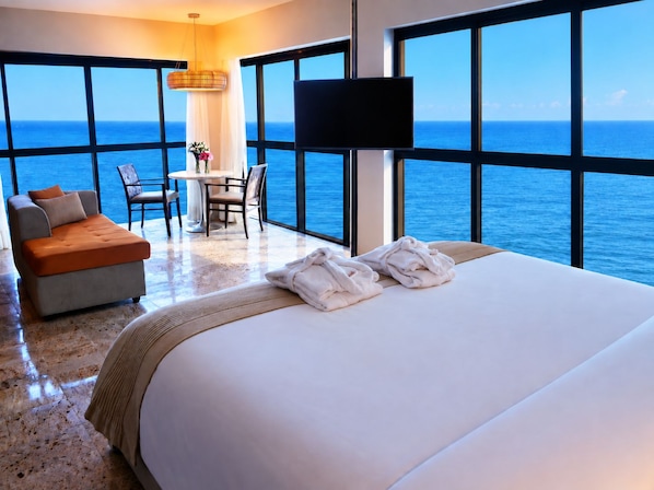 Habitación de hotel con una cama grande, un sofá, una mesa de comedor, sillas y vistas al océano.