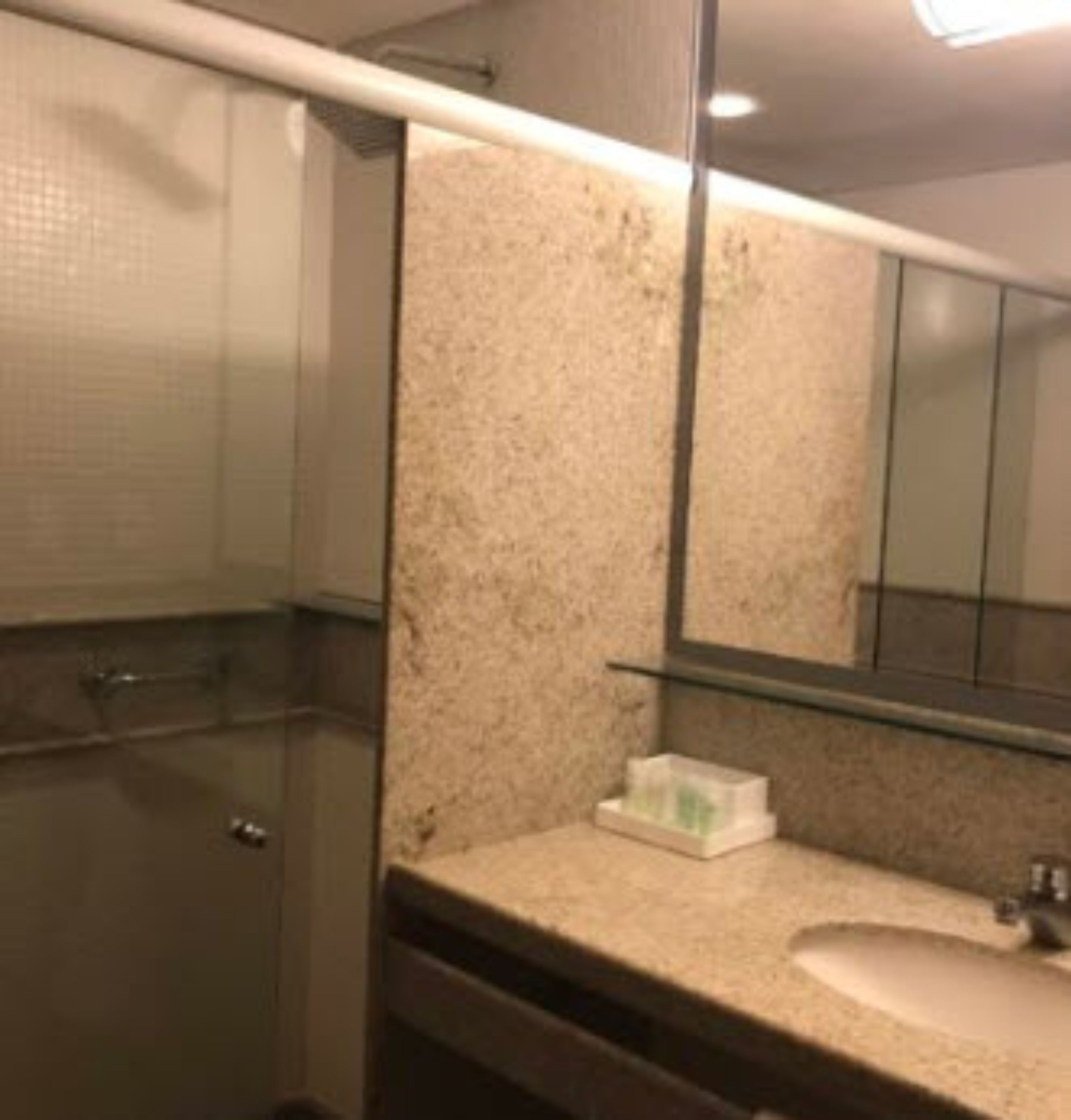 Un baño con cabina de ducha de vidrio, encimera de granito color beige y espejo sobre el lavamanos.