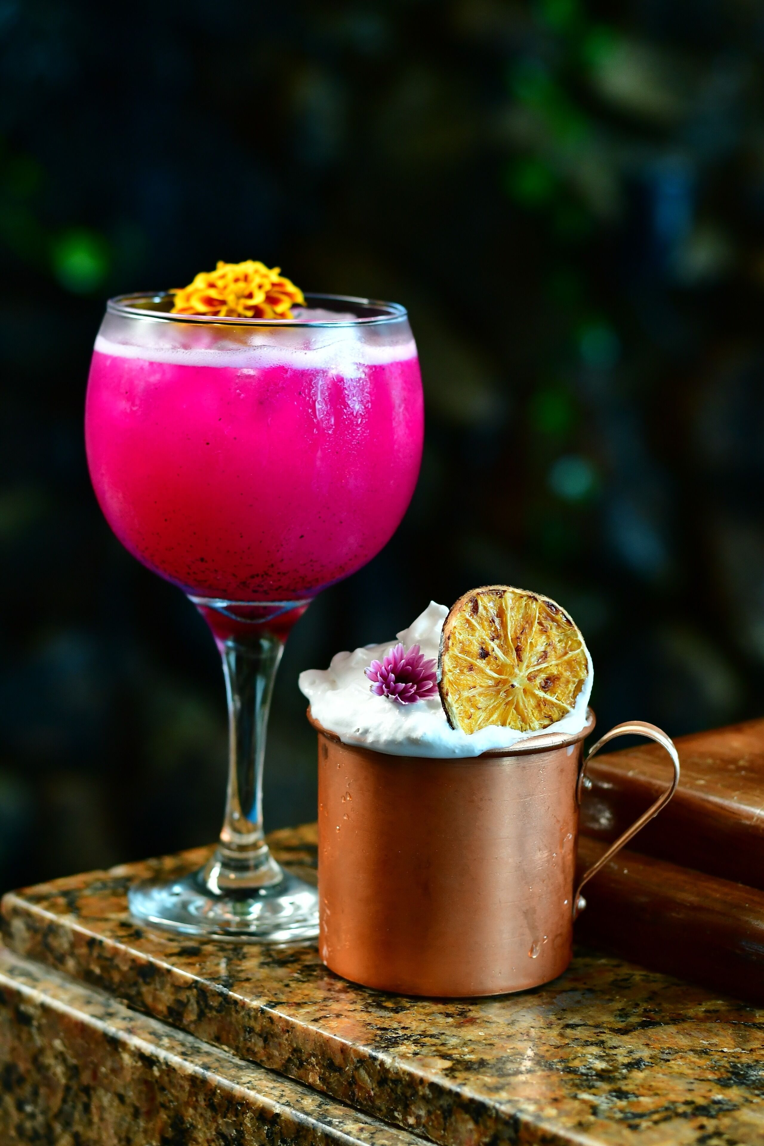 Een cocktail in een glas en een koperen mok met een gedroogde oranje plak en een bloem op een granieten aanrecht.