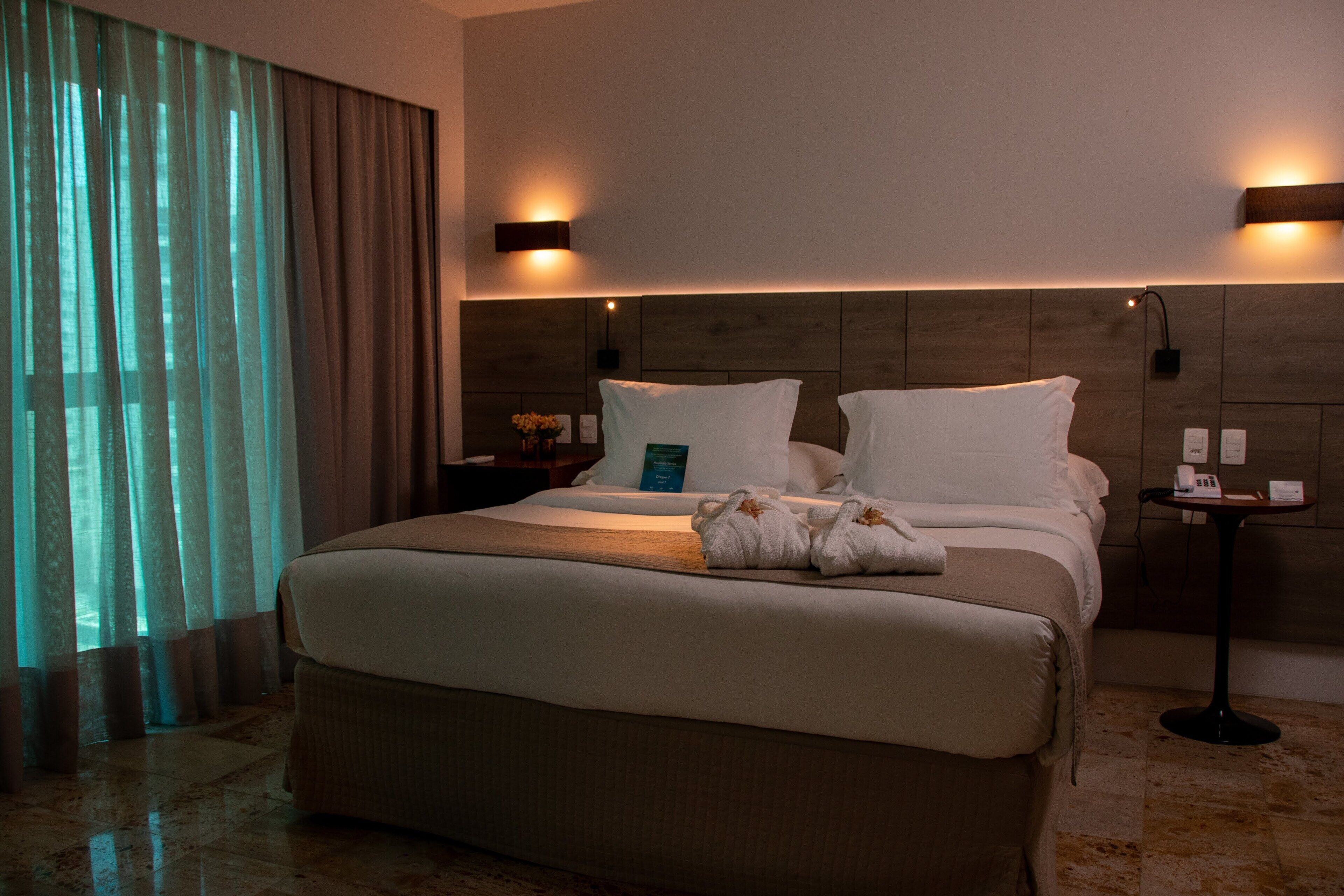 Una habitación de hotel con una cama grande, dos mesitas de noche, una lámpara de pared y un ventanal con cortinas.