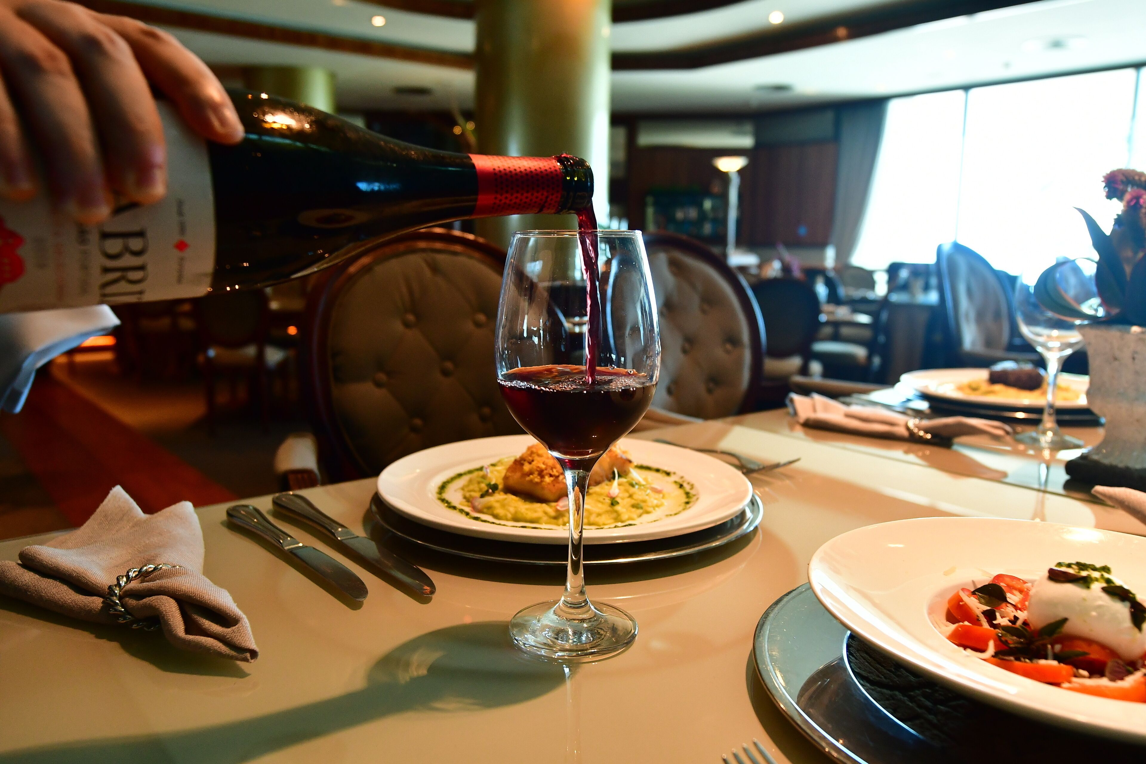 A dining table with a bottle of red wine being poured into a glass, surrounded by plates of food and cutlery.