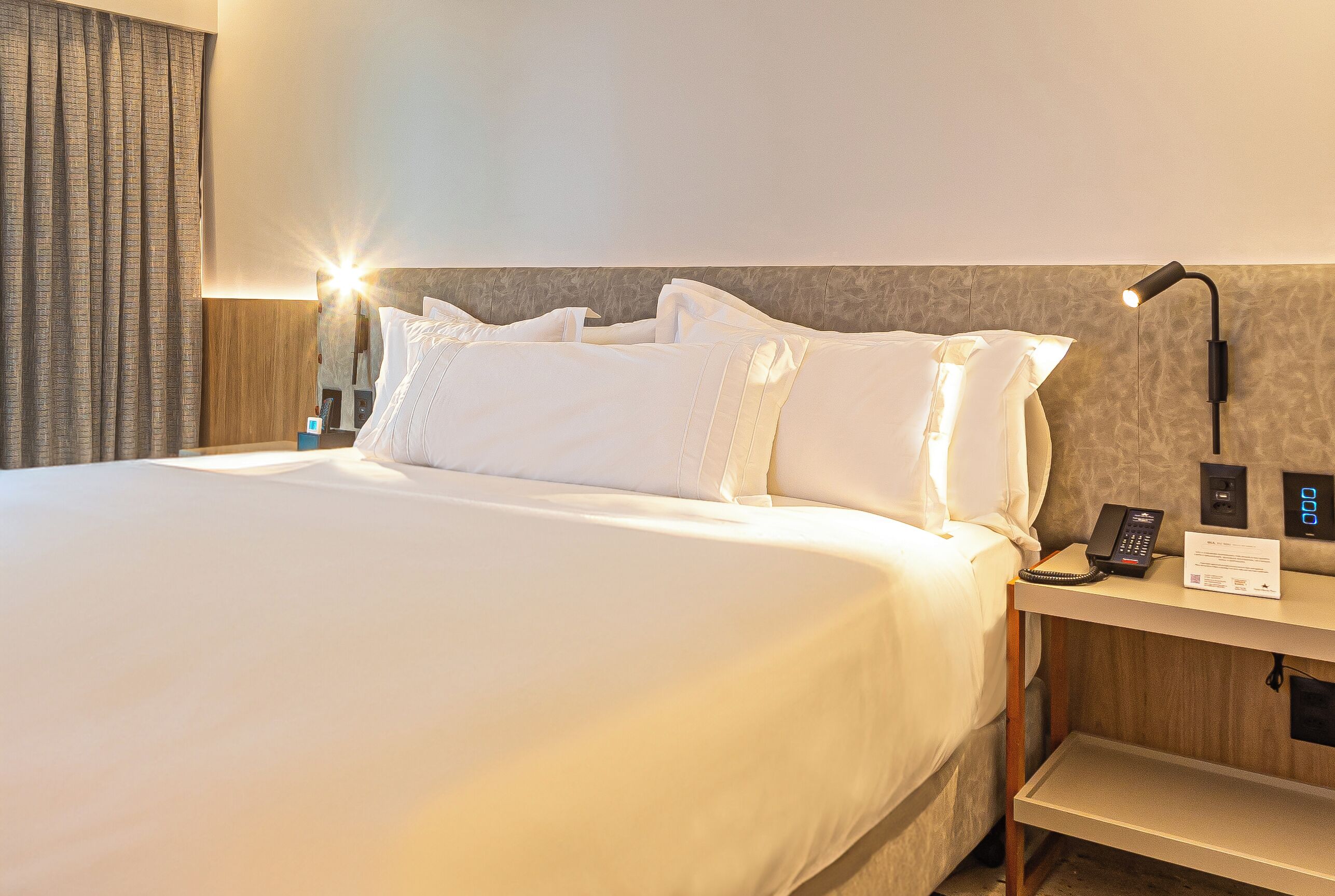 A neatly made bed with white linens, a bedside table with a lamp, a phone, and a sign.