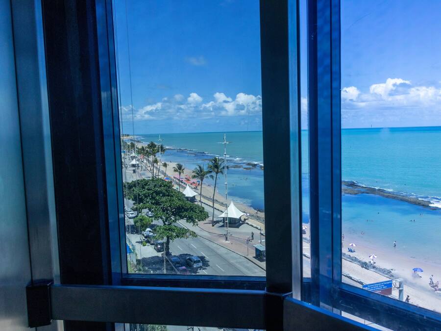 Vista de uma praia pela janela, mostrando o oceano, a praia e uma estrada com palmeiras.