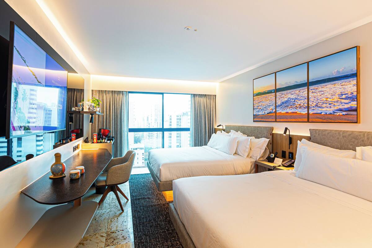 A hotel room with two beds, a desk, a TV, and a large window with a city view.