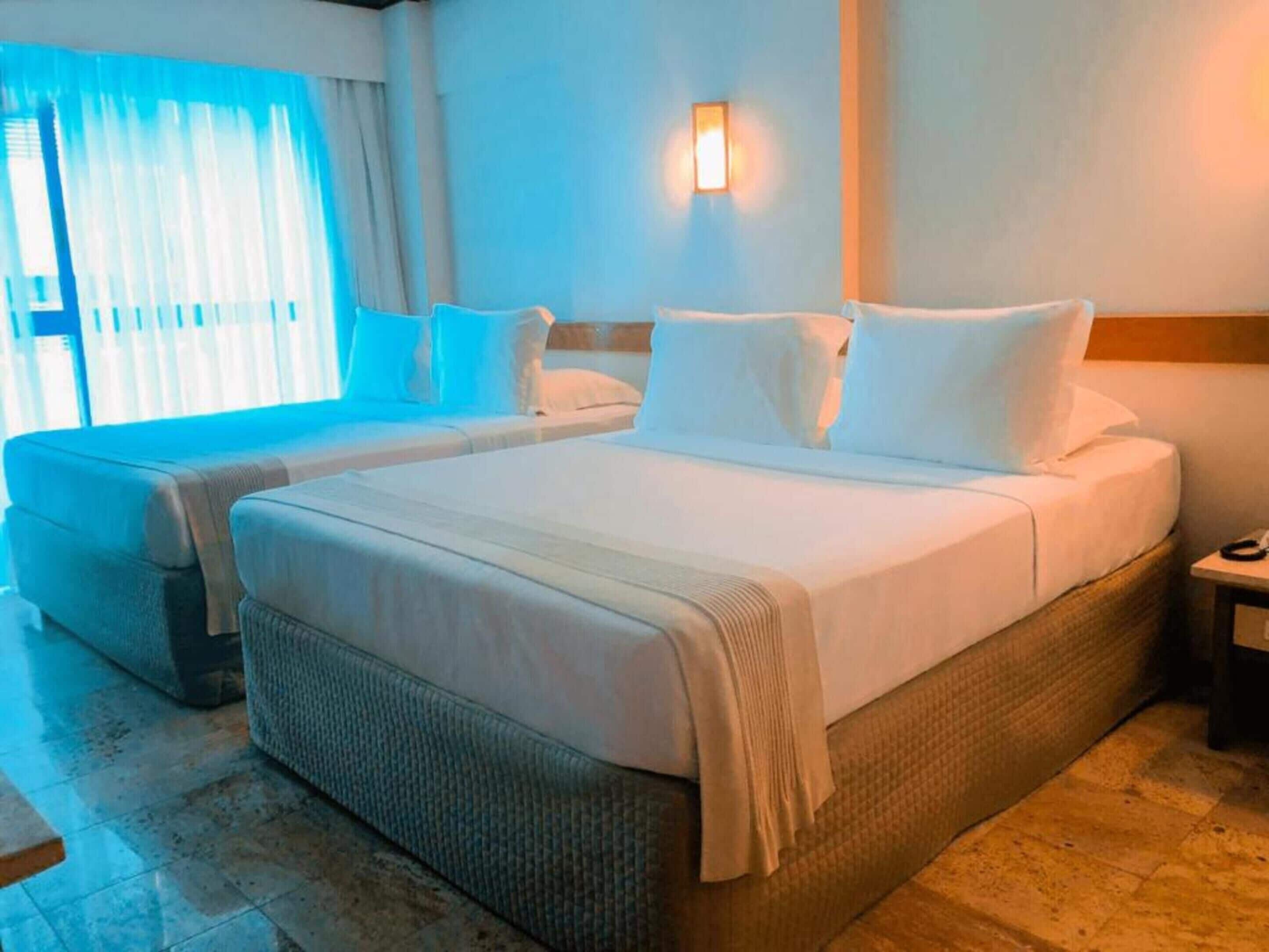 A hotel room with two beds, a large window with curtains, a wall-mounted light, and a bedside table.
