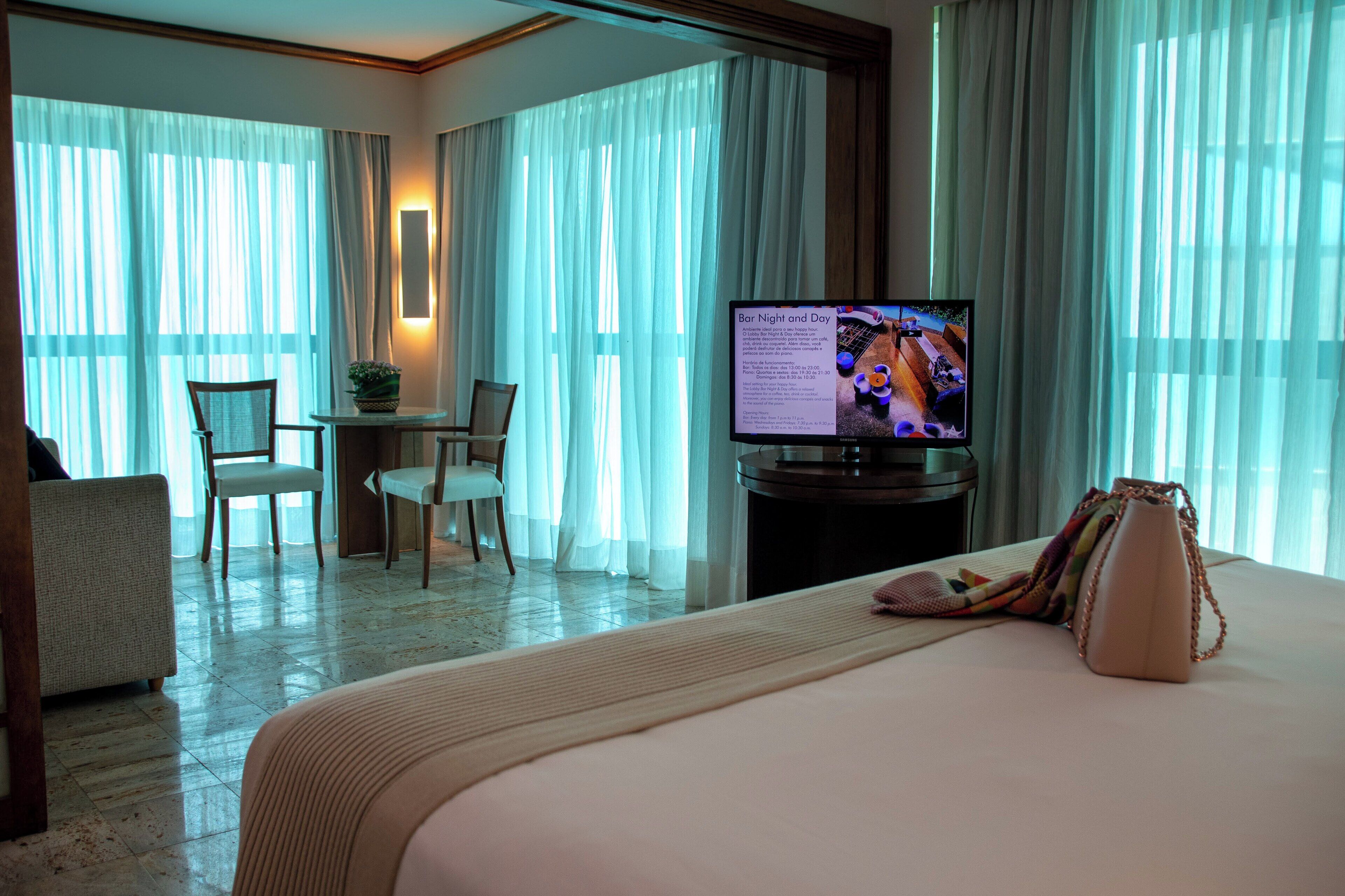Quarto de hotel com uma cama, uma escrivaninha, uma cadeira, uma televisão e vista para o exterior através de cortinas translúcidas.