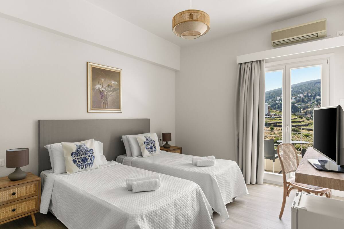 A hotel room with two beds, a desk with a computer, a TV, and a large window with a view.
