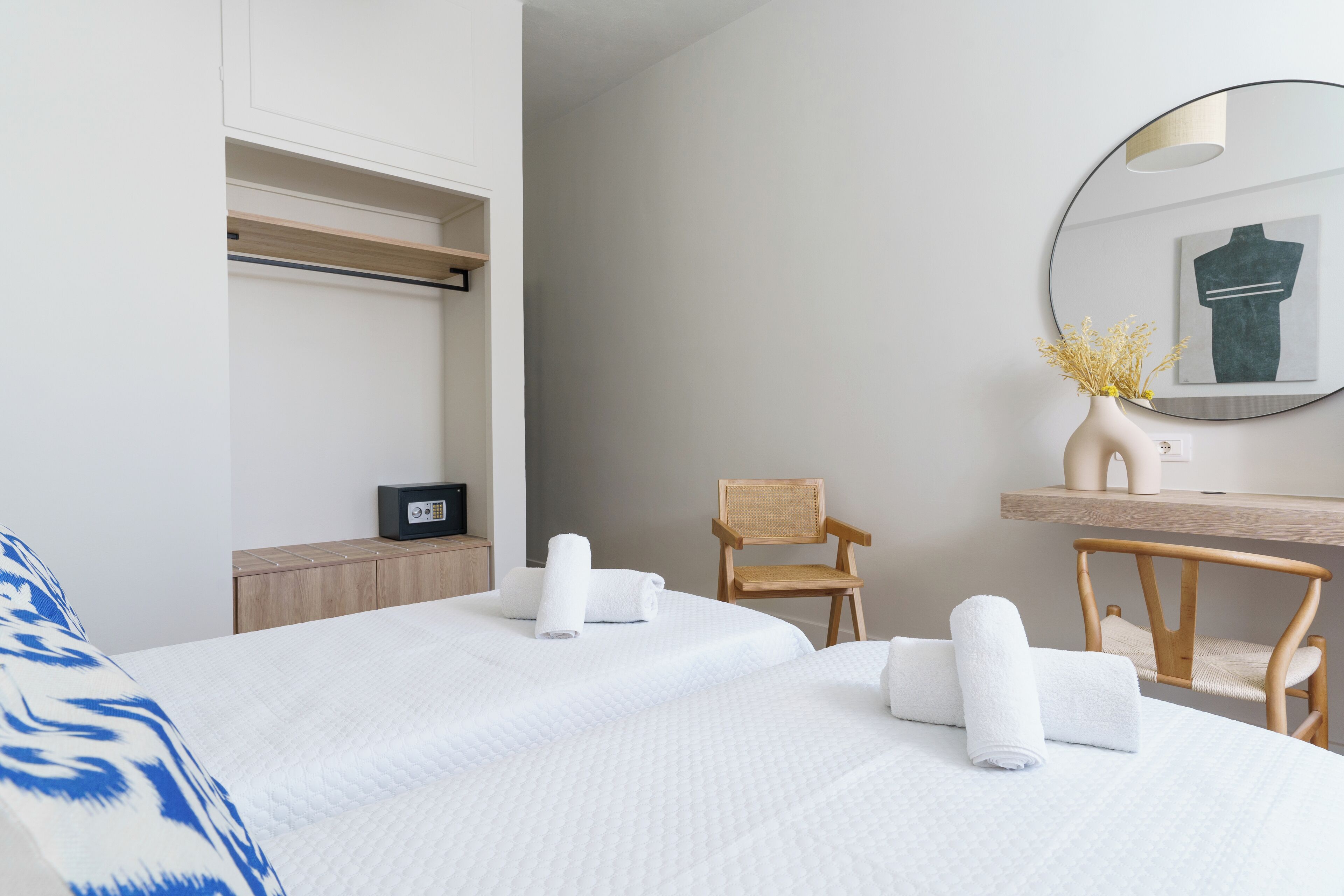 A hotel room with two beds, a wooden chair, a desk with a mirror, and a vase with dried flowers.