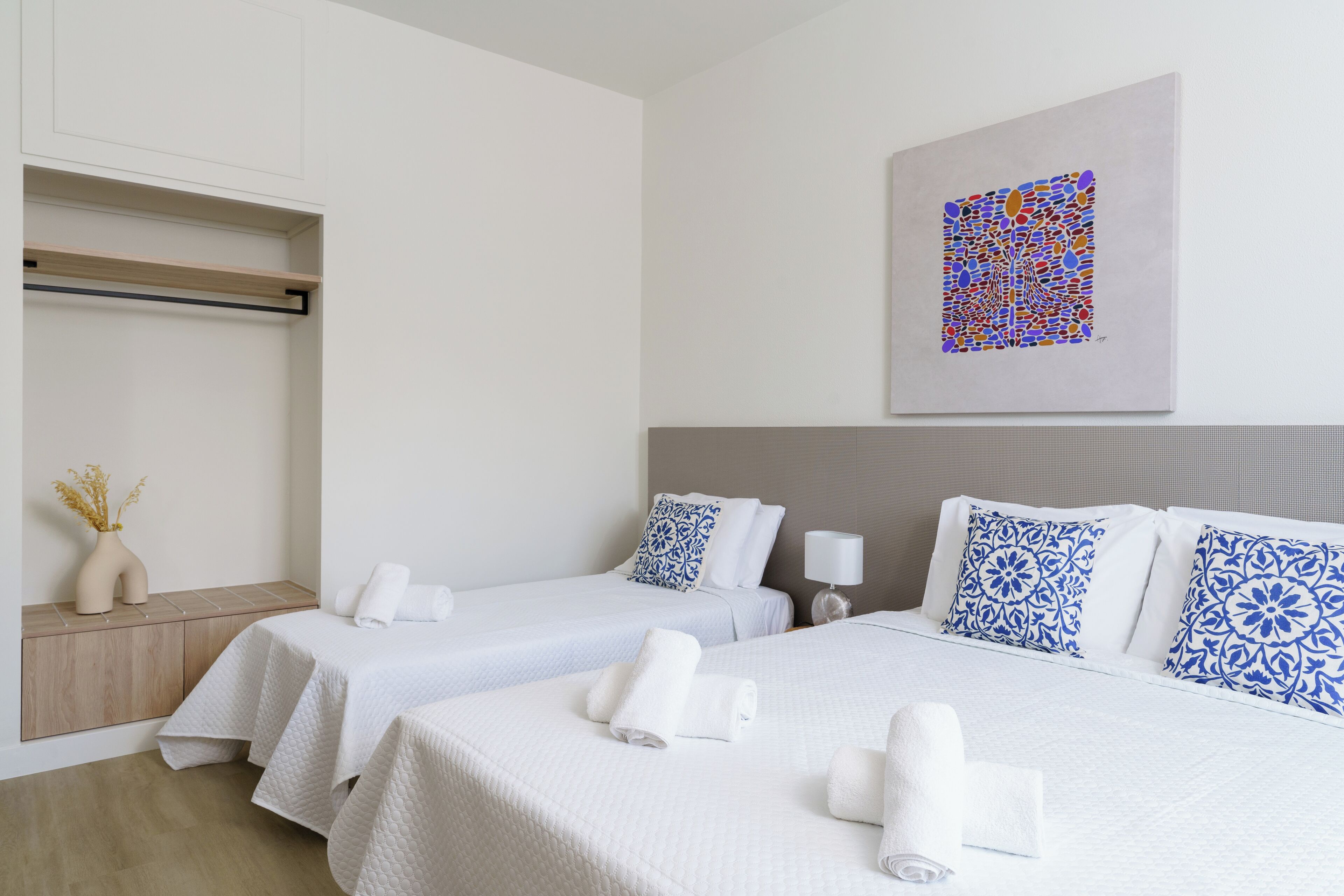 A hotel room with two beds, white bedding, a wall-mounted artwork, a bedside table with a vase, and a wooden cabinet.