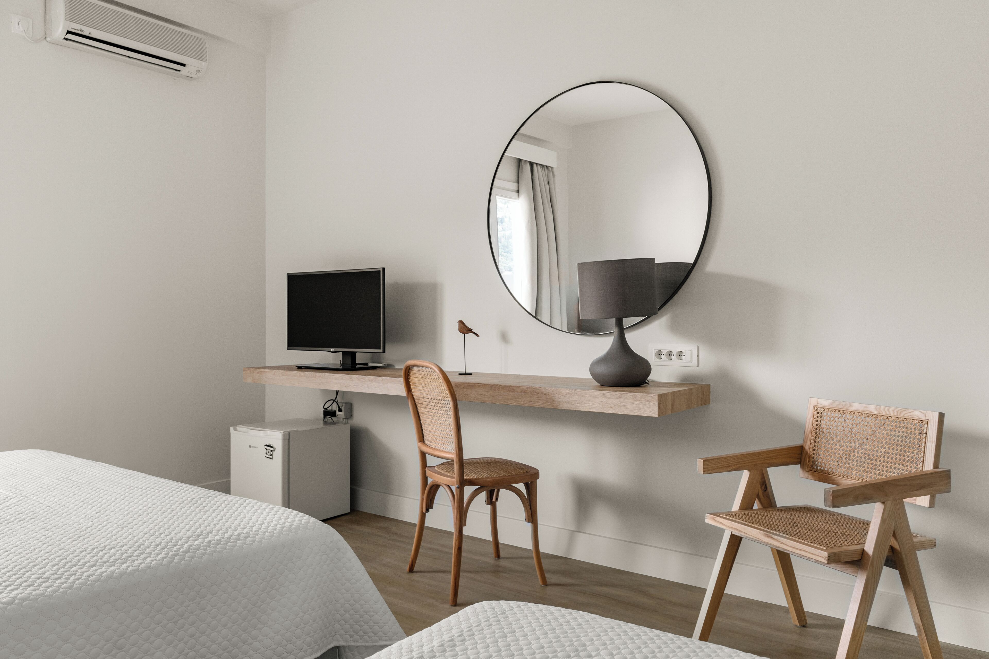 A hotel room with a bed, a desk with a computer, a chair, a round mirror, and a small refrigerator.