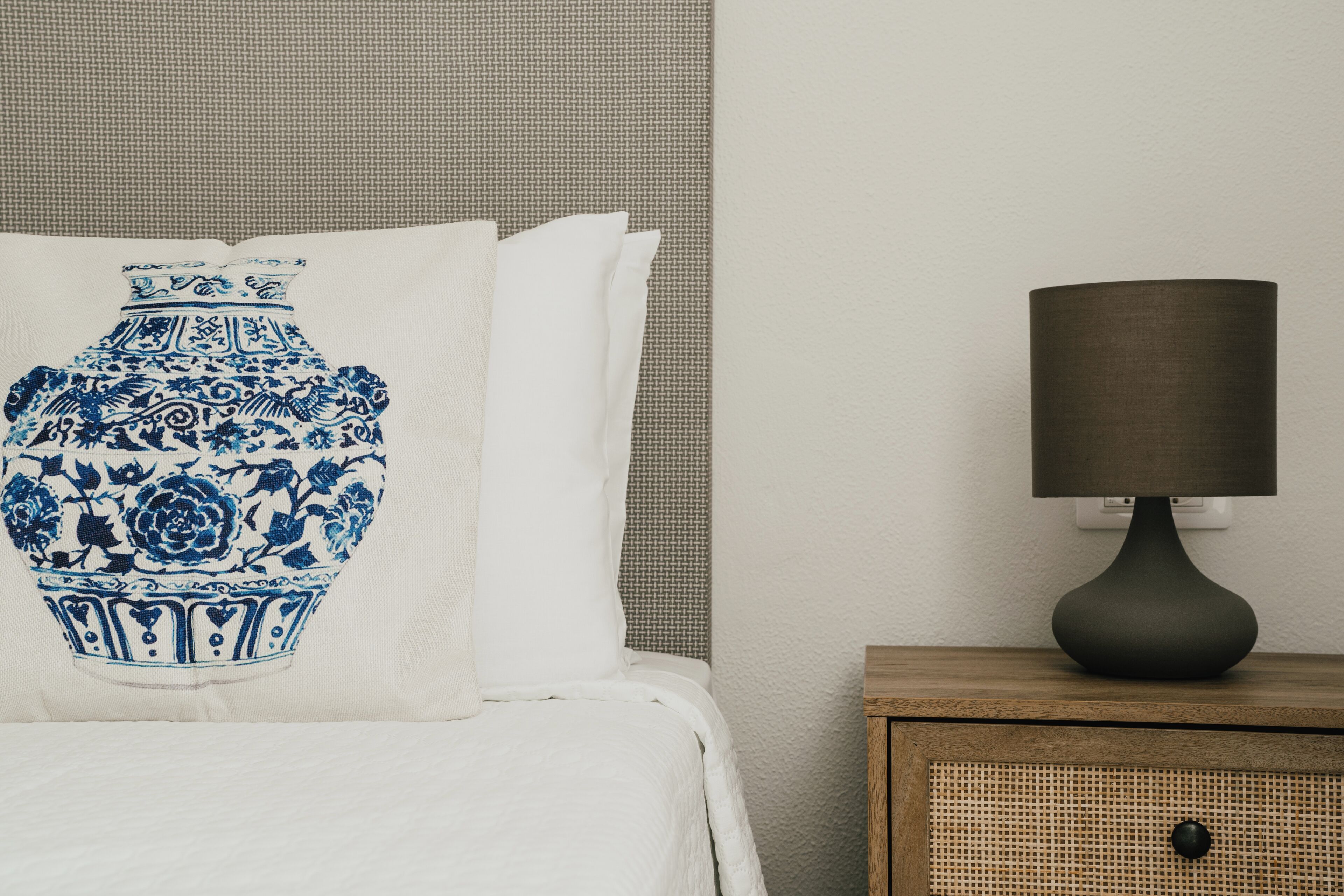 A bed with a white sheet and a pillow with a blue vase design, a dark lamp on a wooden nightstand, and a textured wall.