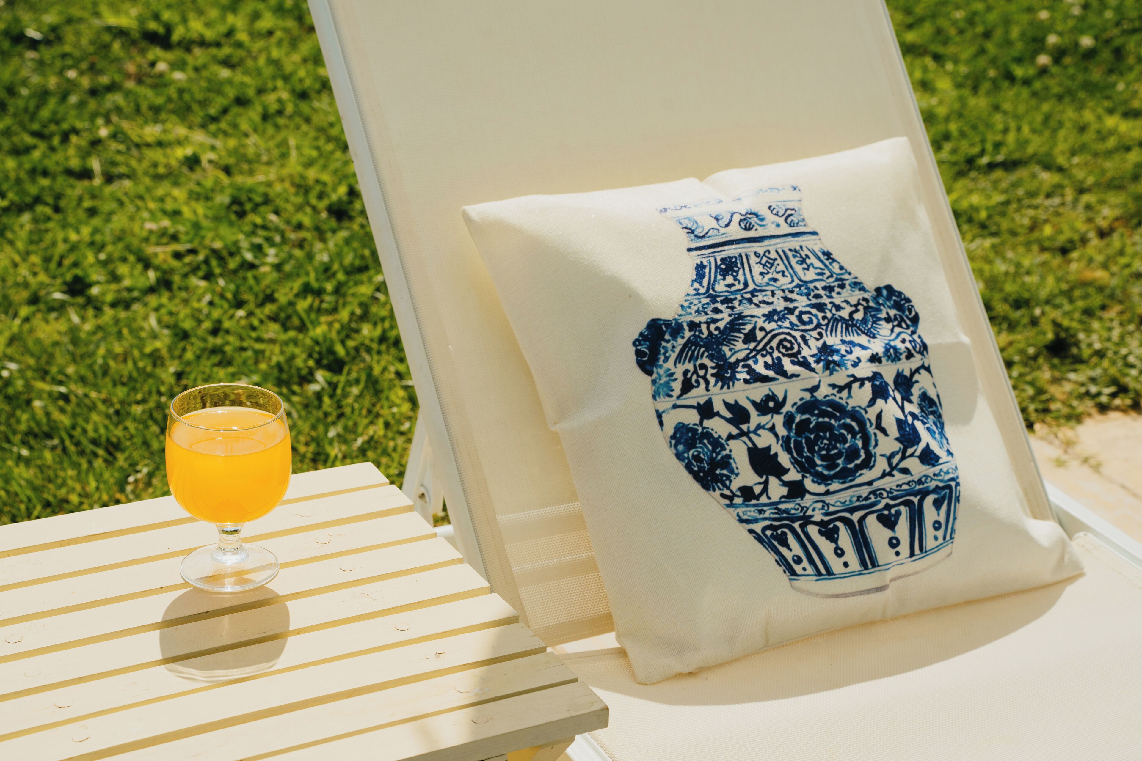 A white lounge chair with a cushion featuring a blue and white floral design, a glass of orange juice, and a grassy background.