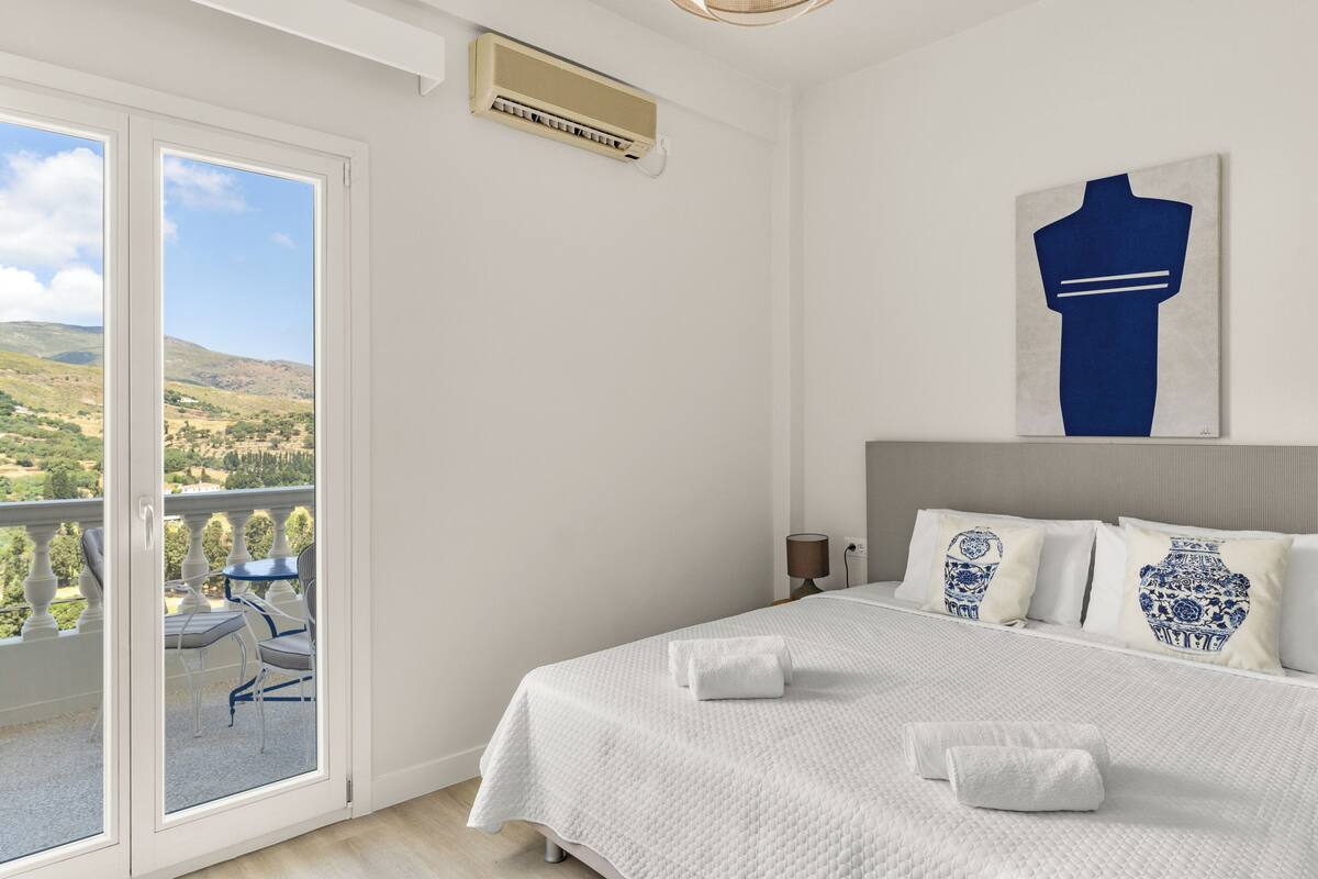 A hotel room with a bed, a balcony with a table and chairs, and a painting on the wall.