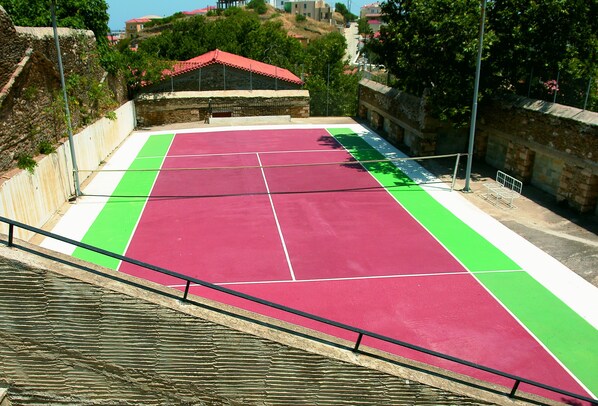 Una cancha de tenis con superficie roja y verde, rodeada de muros de piedra y un edificio con techo rojo.